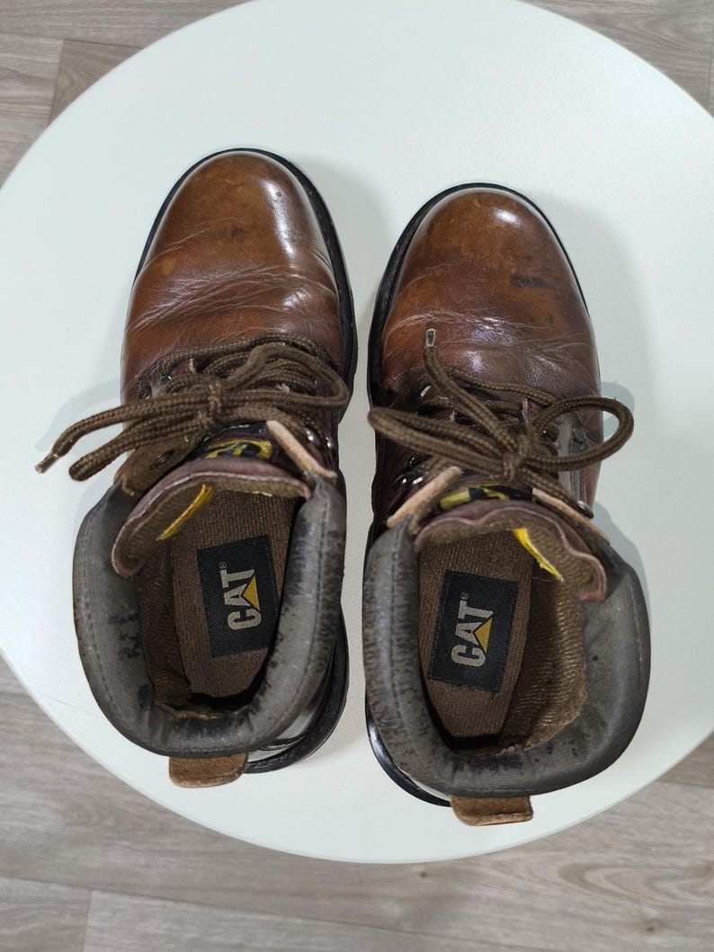 May include: A pair of brown leather work boots with laces, viewed from above. The boots have a worn appearance, with the brand name "CAT" visible inside. The boots are placed on a white surface.