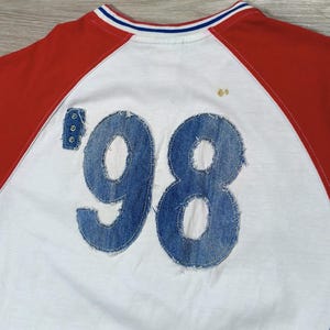 May include: White raglan-sleeve shirt with red sleeves and a blue and white striped collar. The number '98' is appliqued on the back in denim. Three small rivets are visible on the left side of the number.