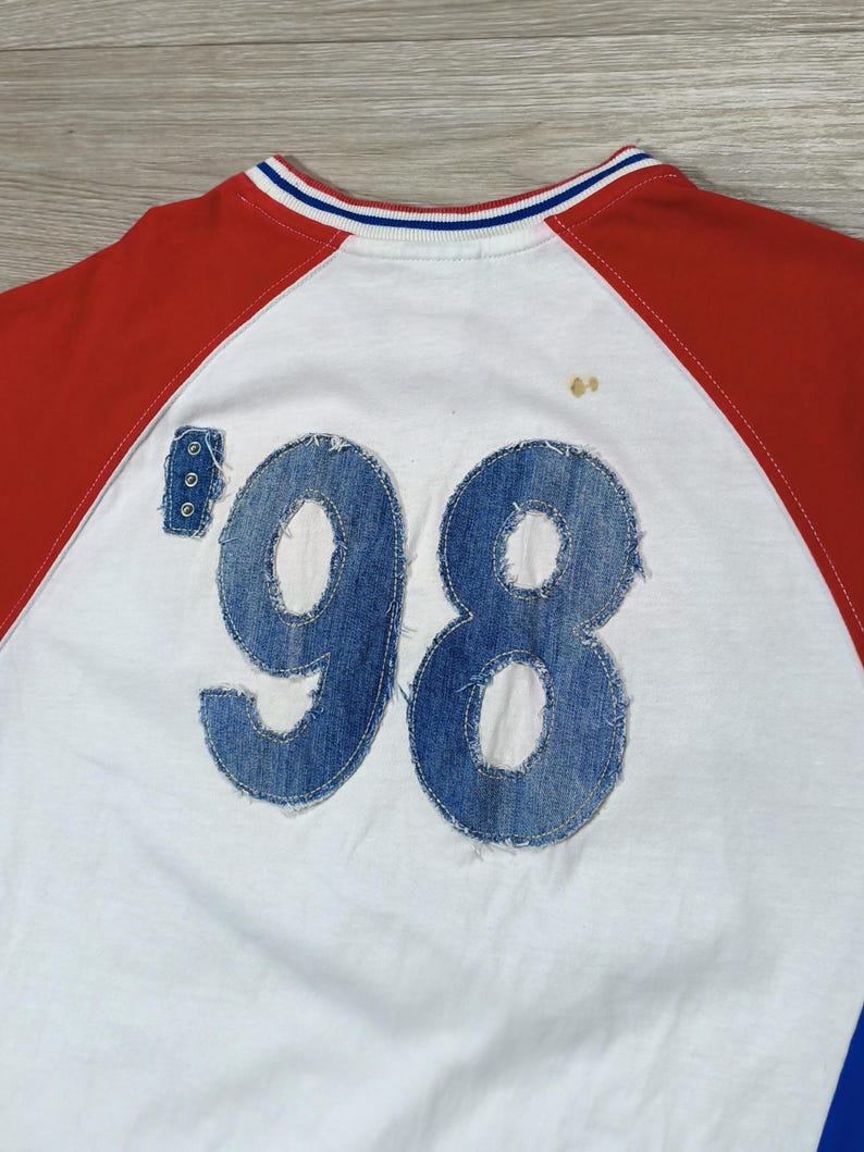 May include: White raglan-sleeve shirt with red sleeves and a blue and white striped collar. The number '98' is appliqued on the back in denim. Three small rivets are visible on the left side of the number.