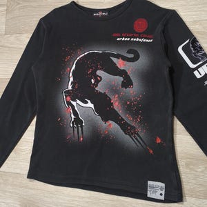 May include: Black long-sleeve shirt featuring a stylized panther graphic in red and white. The shirt has the text "BE SOME ONE urban substance" and a logo on the sleeve. The shirt is made of a soft material.