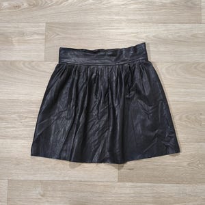 May include: A black faux leather skirt featuring a high waistband and a pleated design. The skirt has a flared silhouette that falls above the knee, with a smooth texture and a visible zipper closure located at the center of the back waistband.