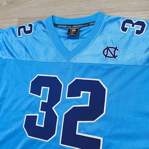May include: Light blue football jersey with the number 32 in navy blue with a white outline. The jersey also features the numbers 2 and 3 on the shoulders, and a logo on the chest. The jersey has a V-neck collar.