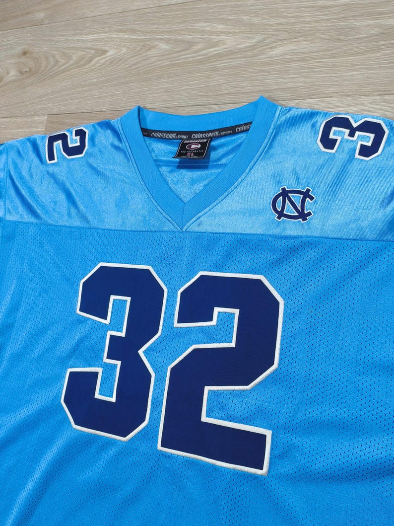 May include: Light blue football jersey with the number 32 in navy blue with a white outline. The jersey also features the numbers 2 and 3 on the shoulders, and a logo on the chest. The jersey has a V-neck collar.