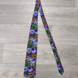 May include: A necktie featuring a dark navy blue background with a vibrant, multi-coloured abstract pattern. The design includes geometric shapes and stylised forms in shades of bright green, purple, light blue, and gold, arranged in a repeating motif.