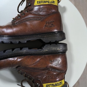 May include: Brown leather work boots with black soles. The boots have yellow and black accents with the brand name "CATERPILLAR" and "STEEL TOE" printed on the side. The boots are laced up with brown laces.