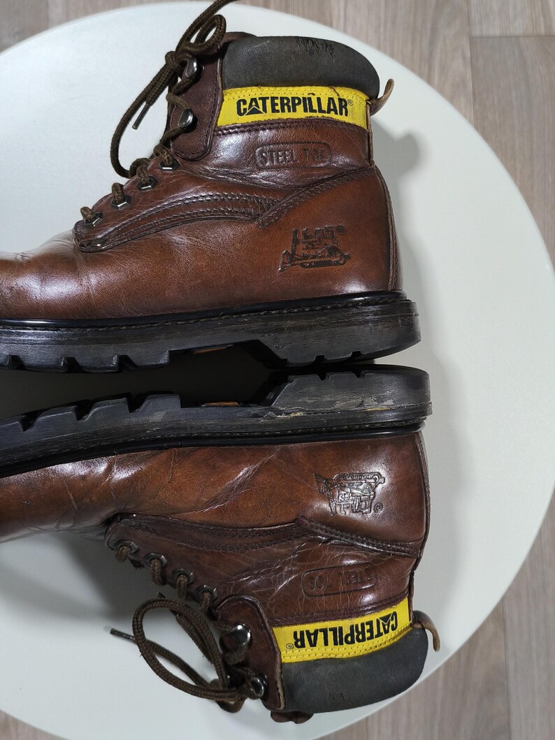 May include: Brown leather work boots with black soles. The boots have yellow and black accents with the brand name "CATERPILLAR" and "STEEL TOE" printed on the side. The boots are laced up with brown laces.