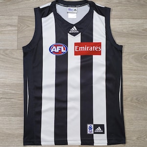 Peut inclure: Un maillot de sport sans manches à rayures verticales noires et blanches. La poitrine présente un logo Adidas, un emblème AFL et un écusson rectangulaire rouge avec le texte Emirates. Un petit logo AFL et la marque Adidas figurent en bas.