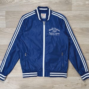 May include: A navy blue bomber jacket with white stripes on the sleeves and trim. The jacket has a white zipper and the text "Rivaldi Premium 1984 Registered Company" on the chest.