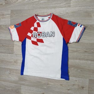 May include: White t-shirt with red sleeves and blue side panels. The shirt features the word "BOBAN" in large letters across the chest, with a checkered design. The sleeves have blue and white trim.