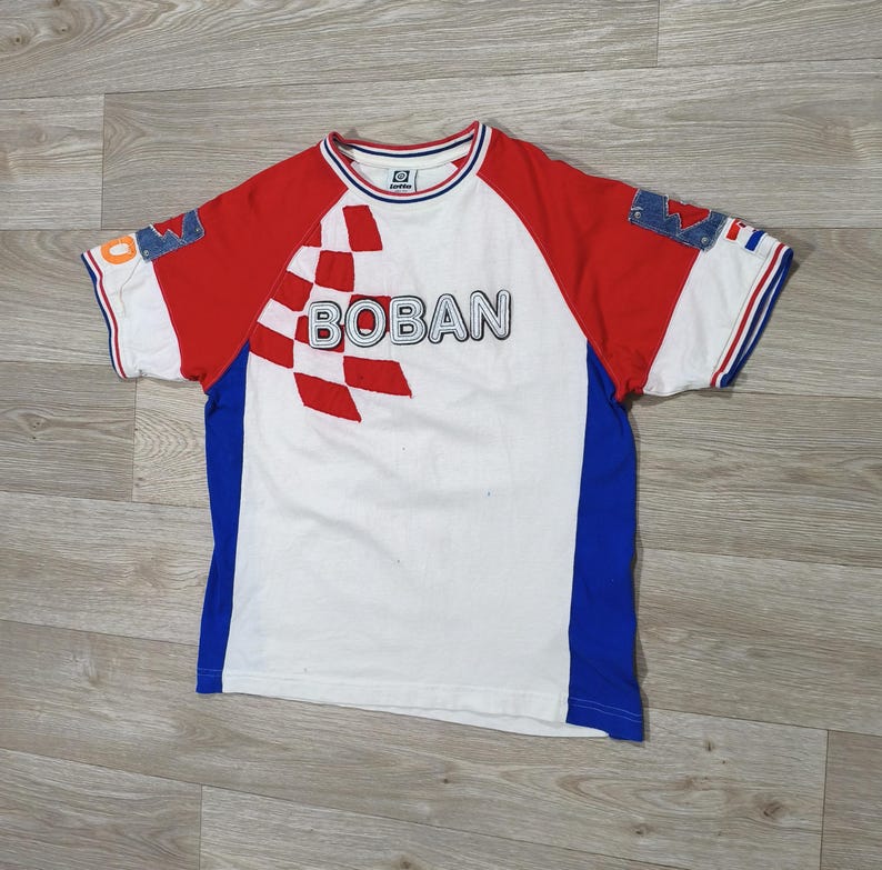 May include: White t-shirt with red sleeves and blue side panels. The shirt features the word "BOBAN" in large letters across the chest, with a checkered design. The sleeves have blue and white trim.
