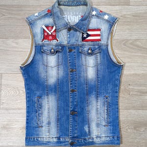 Peut inclure: Un gilet sans manches en jean bleu avec une fermeture boutonnée, deux poches de poitrine et deux poches latérales. Le gilet comporte des écussons avec des drapeaux et des étoiles. Le mot "WAR" est imprimé sur le col.