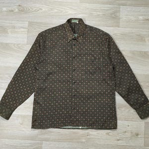 Peut inclure: Une chemise à manches longues avec un motif répété de petits cercles bruns et rouges sur un fond vert foncé. La chemise a un col, une poche poitrine et des poignets boutonnés. La chemise est posée à plat sur une surface en bois clair.