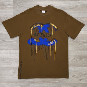 Peut inclure: Un t-shirt marron avec un motif graphique bleu et jaune et le texte "AK-BULLE". Le motif présente un effet dégoulinant et des détails cousus. Le t-shirt a des manches courtes et un col rond.