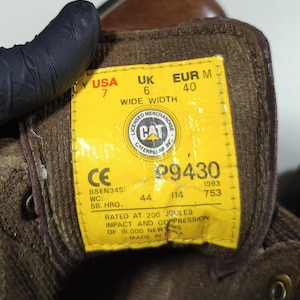 May include: Close-up of a yellow tag inside a brown work boot. The tag displays the brand "CAT" and size information: USA 7, UK 6, EUR M 40. It also includes safety ratings and manufacturing details.