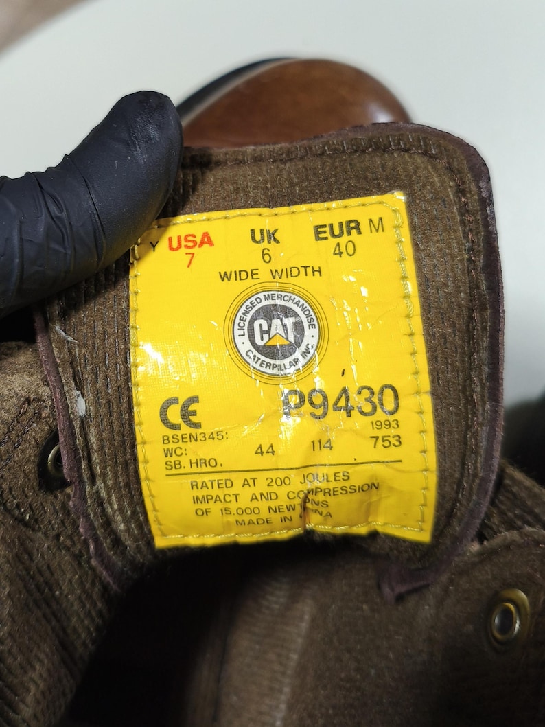 May include: Close-up of a yellow tag inside a brown work boot. The tag displays the brand "CAT" and size information: USA 7, UK 6, EUR M 40. It also includes safety ratings and manufacturing details.