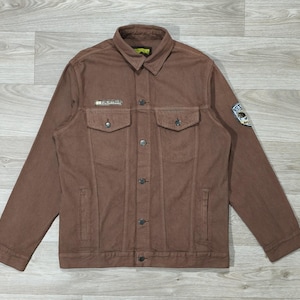 Peut inclure: Veste en jean marron avec fermeture boutonnée, deux poches poitrine et deux poches latérales. La veste a un col et un petit écusson sur le haut de la poitrine gauche. Un écusson circulaire est sur la manche droite.