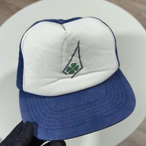 Peut inclure: Casquette de camionneur bleue et blanche avec un logo de trèfle à quatre feuilles. La casquette a un panneau avant blanc avec un motif de trèfle vert et gris, une visière bleue et un dos en filet bleu. Style classique.