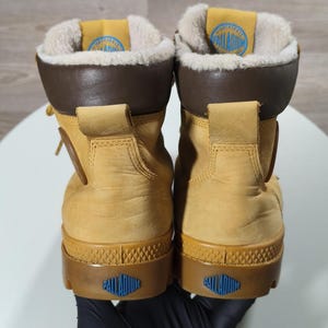 May include: A pair of tan Palladium boots. The boots feature brown collars and soles, with a blue logo on the heel and tongue. The interior is lined with a soft, light-coloured material. These boots are designed for outdoor use.