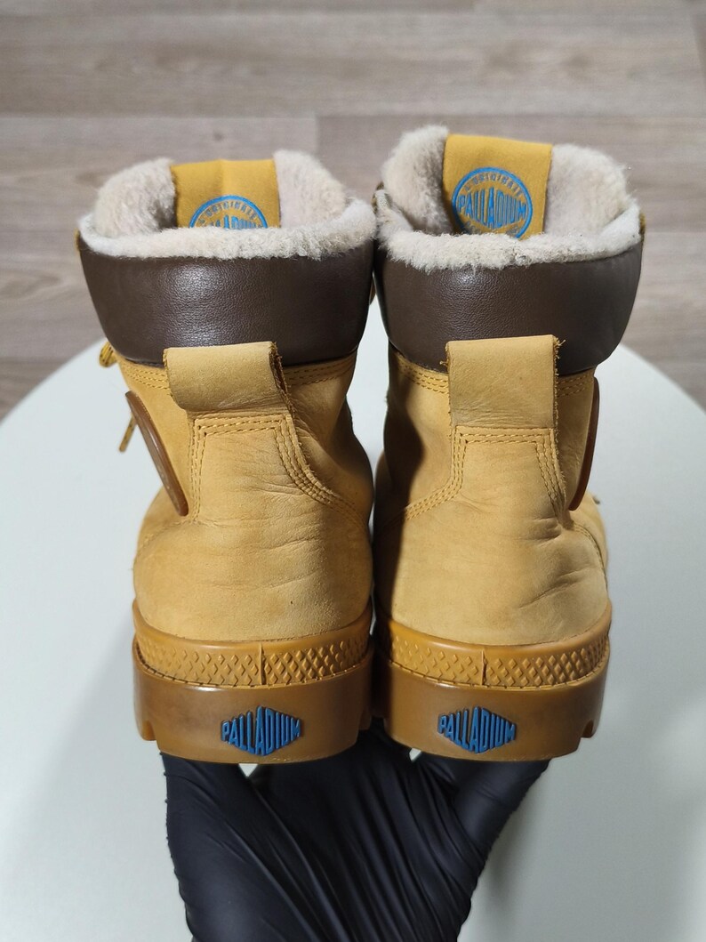 May include: A pair of tan Palladium boots. The boots feature brown collars and soles, with a blue logo on the heel and tongue. The interior is lined with a soft, light-coloured material. These boots are designed for outdoor use.