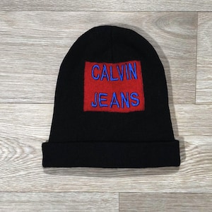 Peut inclure: Bonnet en tricot noir avec un écusson carré rouge brodé des mots "CALVIN JEANS" en bleu. Le bonnet a un bord replié et est présenté sur une surface en bois.