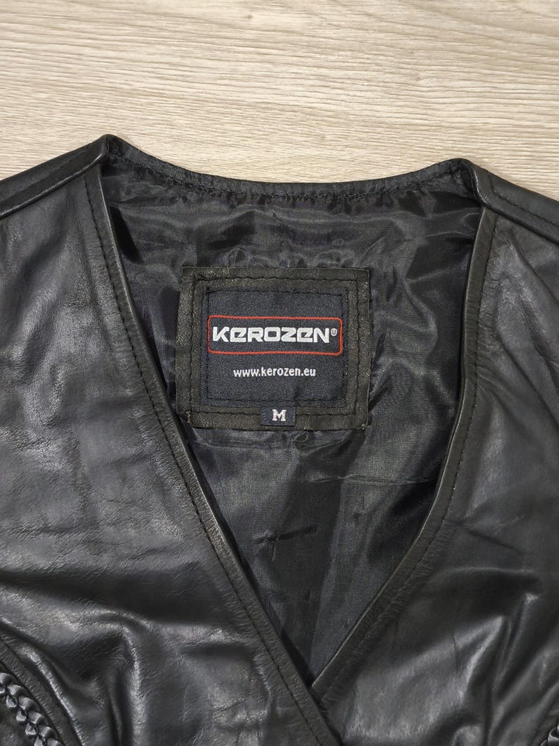 May include: Black leather vest with a V-neck design. The vest features a label with the brand name "Kerozen" and the size "M". The vest is made of a shiny, smooth material.