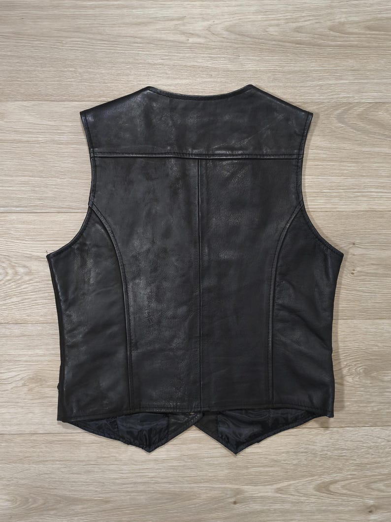 May include: A black leather vest with a classic design. The vest features a V-neck, a tailored fit, and a smooth, shiny finish. The back of the vest is visible, showcasing the stitching and panel details. The vest is likely made of genuine leather.