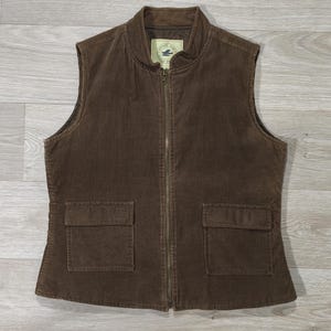 Peut inclure: Un gilet en velours côtelé marron avec une fermeture éclair sur toute la longueur et deux poches avant. Le gilet a un col montant et une petite étiquette près du col. Le gilet est en tissu de velours côtelé côtelé.
