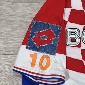 May include: A red, white, and blue soccer jersey with the number 10 in orange. The jersey features a denim patch with a logo and a checkered pattern on the front. The sleeves are red and white.