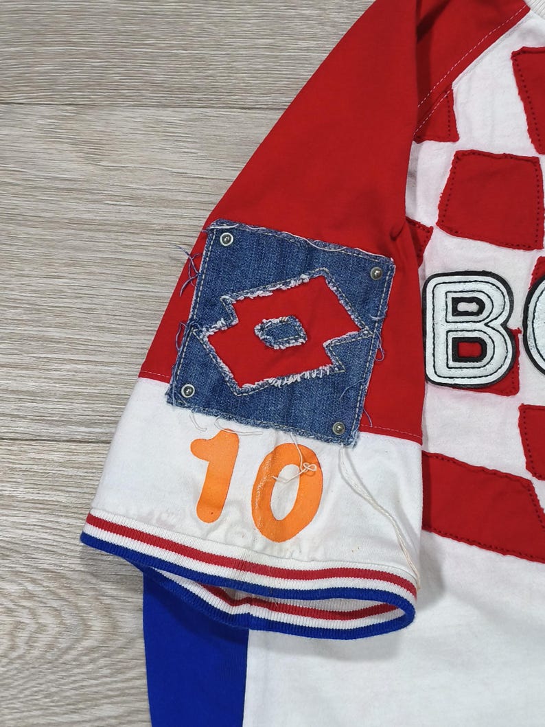 May include: A red, white, and blue soccer jersey with the number 10 in orange. The jersey features a denim patch with a logo and a checkered pattern on the front. The sleeves are red and white.