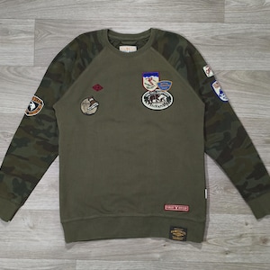 Peut inclure: Sweat-shirt vert olive avec manches imprimées camouflage et plusieurs écussons brodés. Les écussons comportent du texte et divers motifs. Le sweat-shirt a un col rond et des poignets côtelés. La marque "Finest Division" est visible sur un écusson.