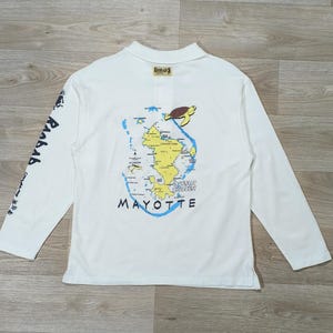 Peut inclure: Un polo blanc à manches longues avec une impression graphique au dos représentant une carte jaune de Mayotte, une tortue de mer brune et des contours côtiers bleus. Le texte MAYOTTE est imprimé en noir sous la carte, avec la marque Raobab sur la manche.