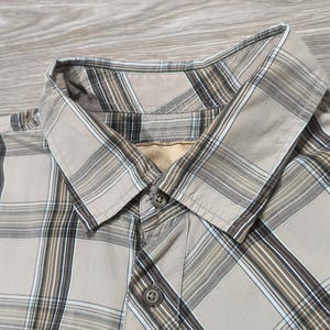 May include: Close-up of a tan plaid button-down shirt. The shirt has a classic collar and two visible buttons. The plaid pattern includes shades of brown, white, and tan, creating a neutral colour palette. The shirt is made of a lightweight fabric.
