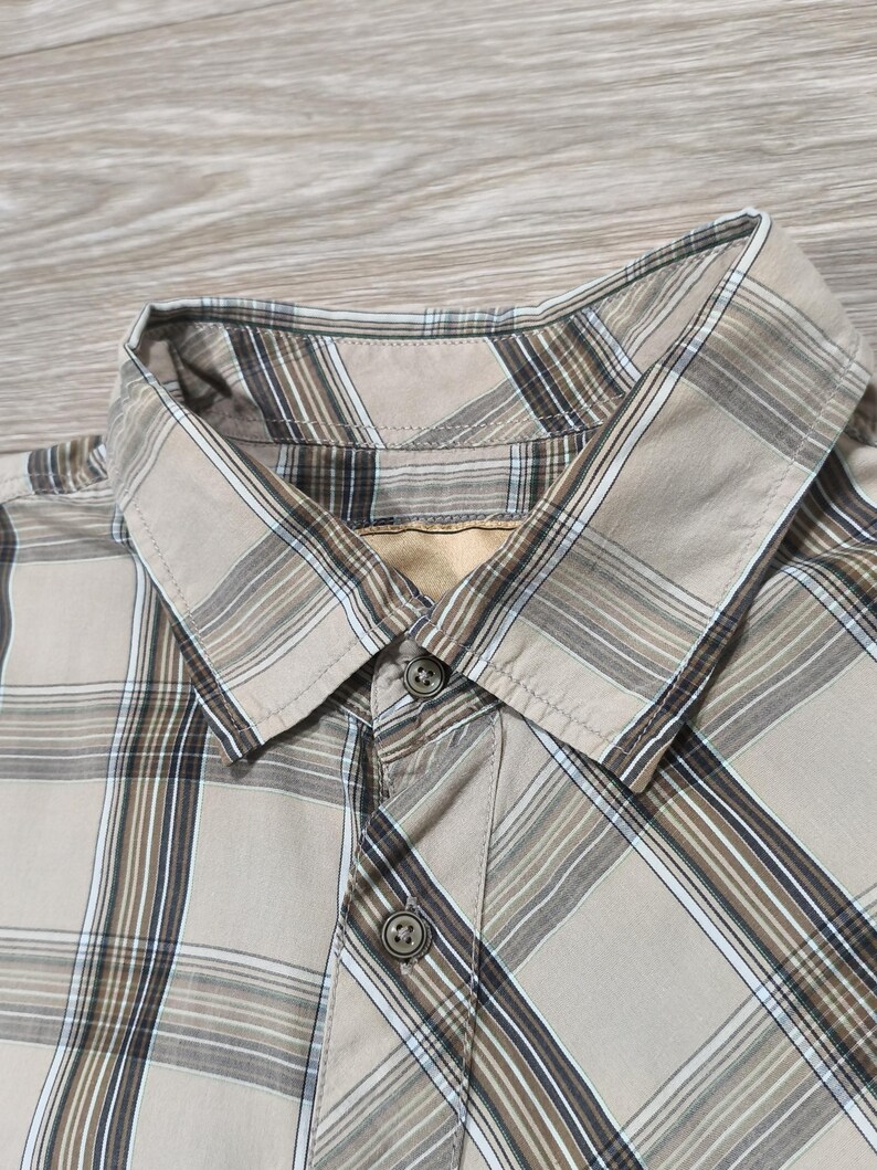 May include: Close-up of a tan plaid button-down shirt. The shirt has a classic collar and two visible buttons. The plaid pattern includes shades of brown, white, and tan, creating a neutral colour palette. The shirt is made of a lightweight fabric.