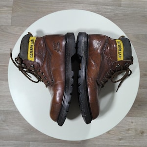 May include: Pair of brown leather work boots with black soles. The boots have a yellow band with the word "CATERPILLAR" on the back. The boots are laced up and appear to be worn.