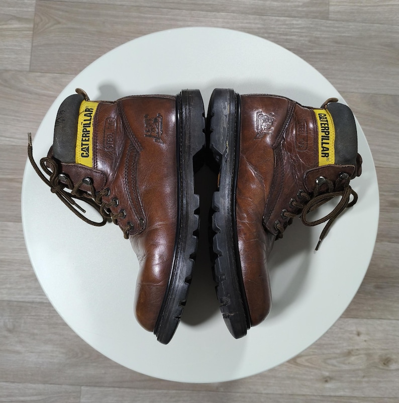 May include: Pair of brown leather work boots with black soles. The boots have a yellow band with the word "CATERPILLAR" on the back. The boots are laced up and appear to be worn.