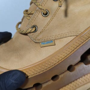May include: Close-up of a tan leather boot with a brown rubber sole. The boot features a lace-up design with metal eyelets and the brand name "PALLADIUM" on a small tag.