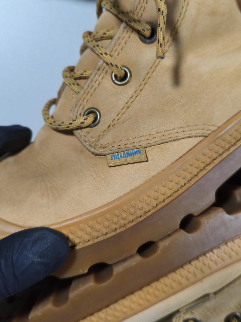 May include: Close-up of a tan leather boot with a brown rubber sole. The boot features a lace-up design with metal eyelets and the brand name "PALLADIUM" on a small tag.