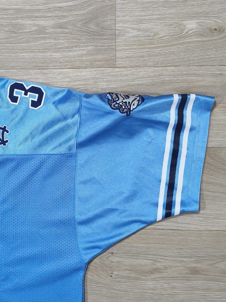 May include: Light blue jersey with the number 3 in navy blue on the shoulder. The sleeve has navy and white stripes. A logo is visible on the shoulder. The jersey is made of a breathable, mesh-like fabric.