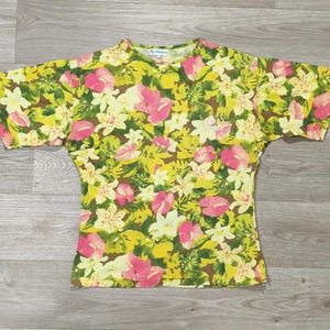 Peut inclure: Un t-shirt à manches courtes avec un motif floral vif. Le t-shirt présente un mélange de fleurs roses, blanches et jaunes sur un fond vert et marron. Le t-shirt est posé à plat sur une surface en bois.