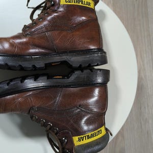 May include: Brown leather work boots with the word "CATERPILLAR" in yellow on the collar. The boots have a lace-up design and a thick black sole. The boots are sitting on a white surface.