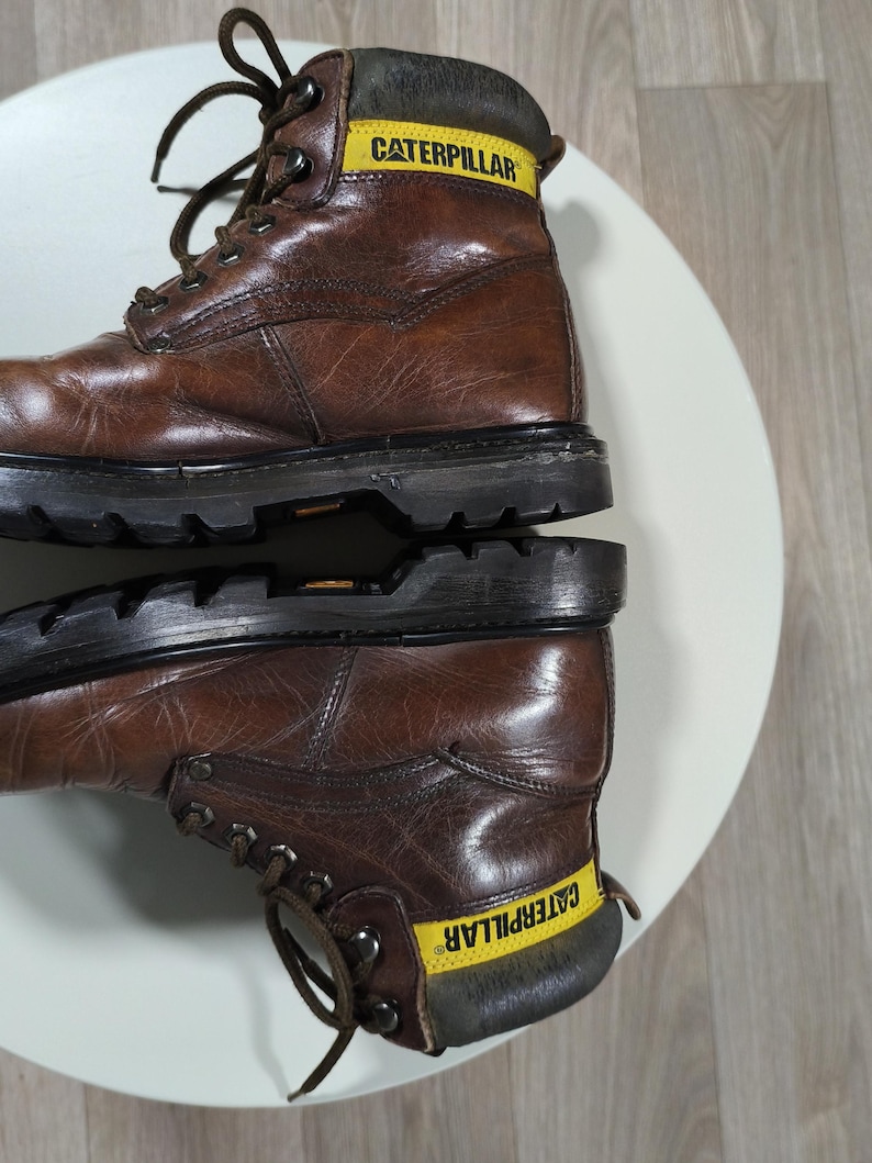 May include: Brown leather work boots with the word "CATERPILLAR" in yellow on the collar. The boots have a lace-up design and a thick black sole. The boots are sitting on a white surface.