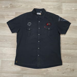 Peut inclure: Chemise bleu marine foncé à manches courtes avec deux poches de poitrine et des écussons brodés. La chemise a un col classique et un petit logo en bas. Fabriquée dans un tissu résistant.