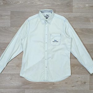 Peut inclure: Une chemise boutonnée bleu clair à manches longues. La chemise présente des coutures bleu clair le long des coutures et une petite poche sur la poitrine avec le texte "Harrisons & Sons". La chemise a un col boutonné.