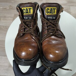 May include: A pair of brown leather work boots with the "CAT" logo in yellow and black on the tongue. The boots have a steel toe and are laced up with brown laces. The soles are black.