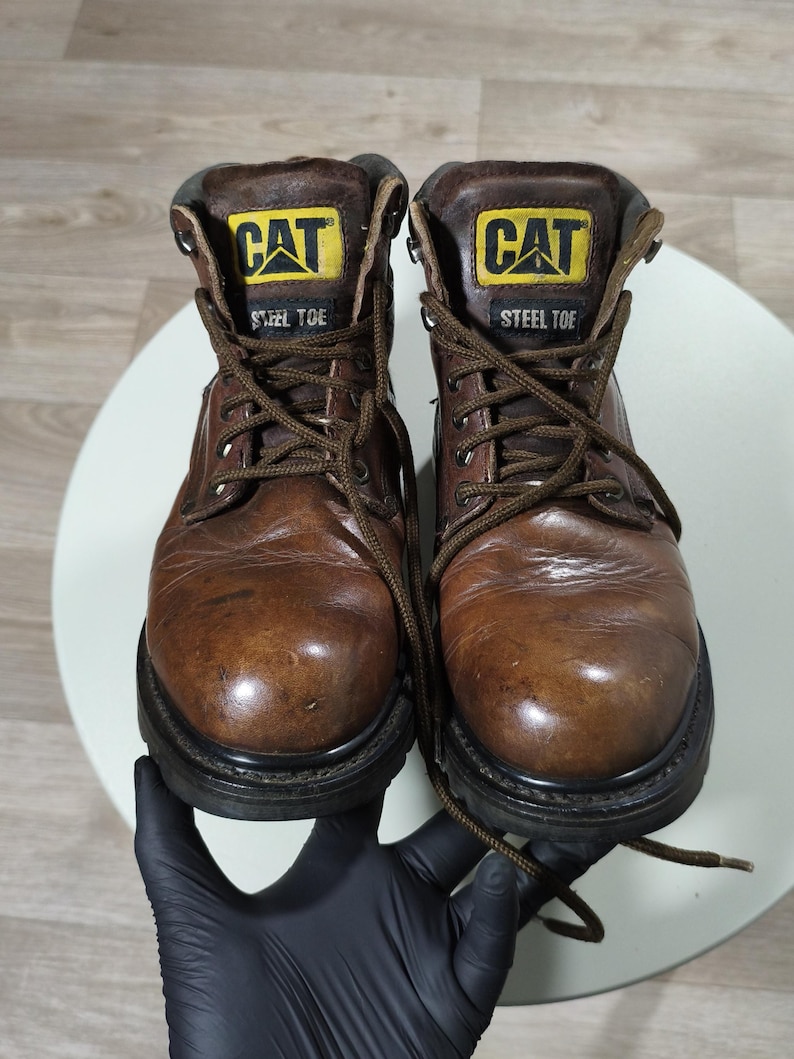 May include: A pair of brown leather work boots with the "CAT" logo in yellow and black on the tongue. The boots have a steel toe and are laced up with brown laces. The soles are black.
