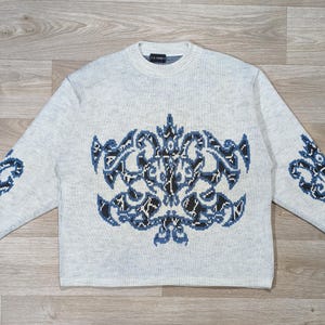 Peut inclure: Un pull gris clair avec un col ras du cou et des manches longues. Le pull présente un grand motif bleu et noir orné sur le devant et des motifs assortis plus petits sur les manches. Le pull est en maille douce.