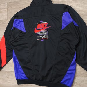 Peut inclure: Veste de survêtement noire avec des détails rouges et violets. Le logo Nike rouge et blanc est visible au dos, avec le texte "ONE BOWERMAN DRIVE". Des rayures rouges ornent les manches.