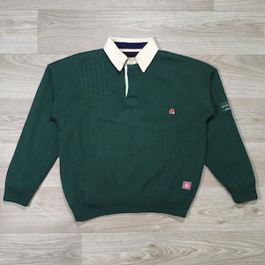 Op de afbeelding: Een donkergroen rugbyshirt met lange mouwen, een crèmekleurige kraag, een knoopsluiting en verticale kabelgebreide texturen op de borst. Het shirt heeft een klein rood geborduurd logo op de borst en een rechthoekige patch op de mouw.