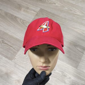 Peut inclure: Une casquette de baseball rouge vif avec un logo brodé sur le devant. Le logo se compose d'une lettre A stylisée en blanc et bleu, avec une tête d'aigle dorée intégrée au motif. La casquette possède une calotte structurée et une visière courbée.
