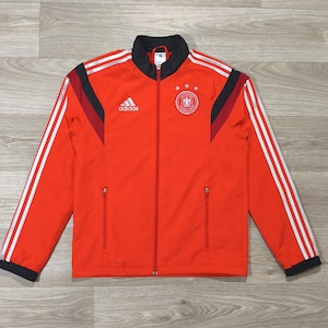 Könnte beinhalten: Rote Adidas Trainingsjacke mit weißen Streifen auf den Ärmeln. Die Jacke hat einen schwarzen Kragen und Akzente, ein deutsches Nationalmannschaftswappen und das Adidas-Logo. Sie hat einen durchgehenden Reißverschluss und zwei Seitentaschen.